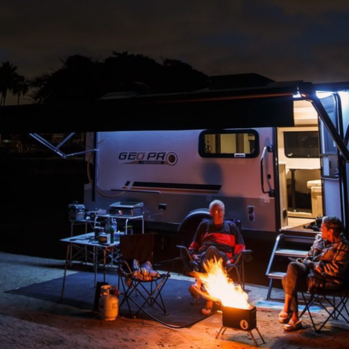 Nighttime lifestyle photo of two people seated by a lit firebox next to a parked camper with awning lights on