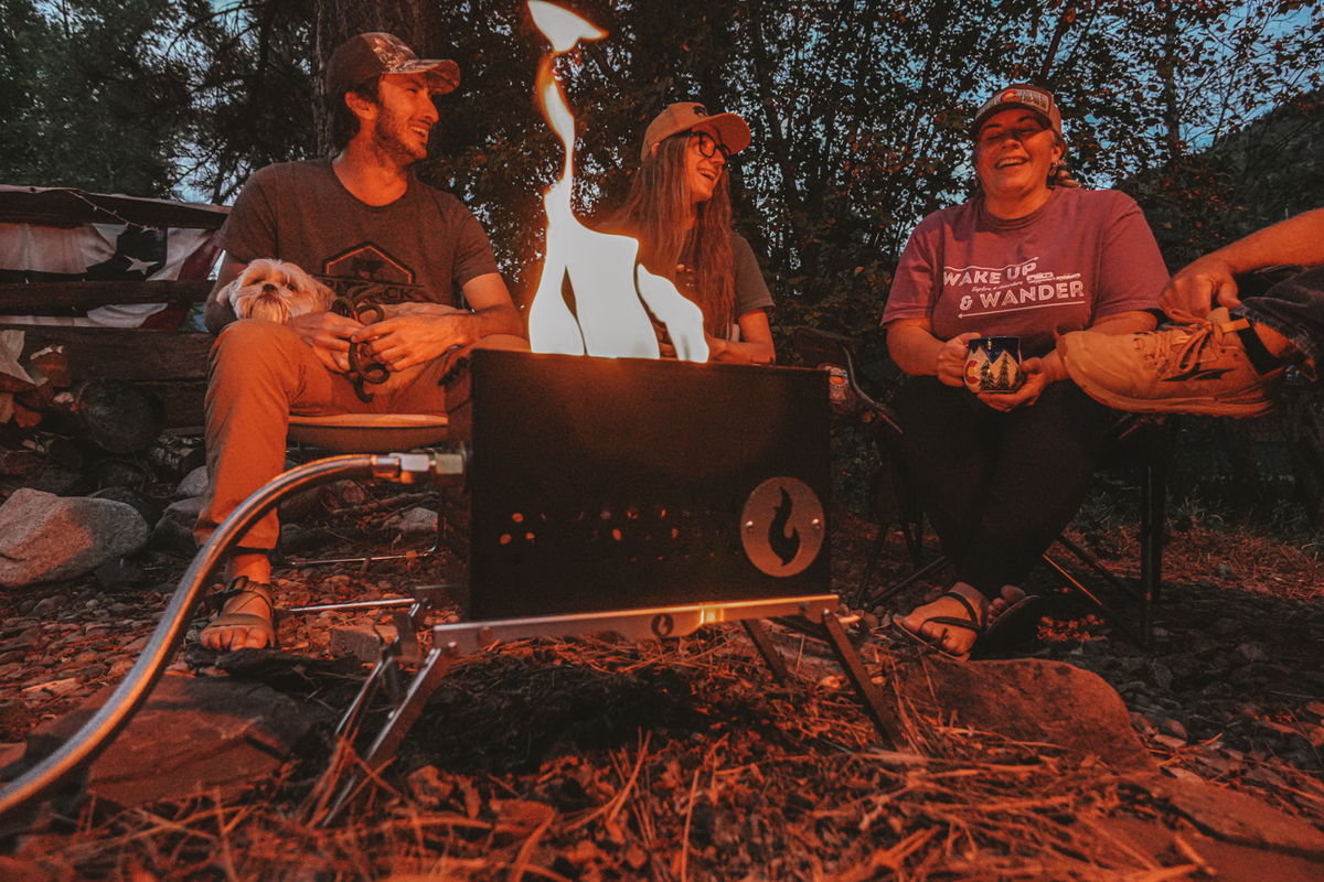 Lifestyle photo of three people relaxing around a lit firebox at a campsite at dusk