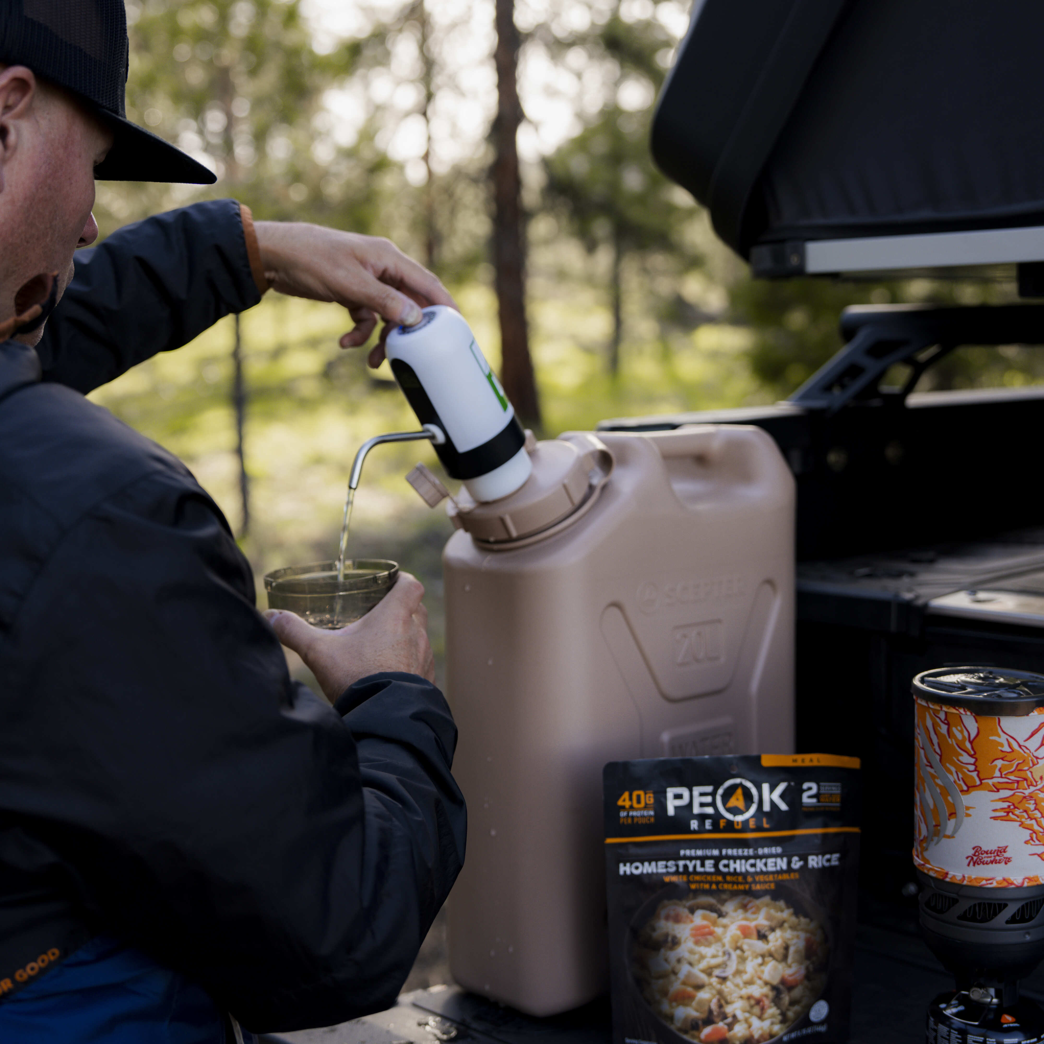 Camper filling a cup from the Lone Rock Concepts pump on a tan 20L Scepter can beside a freeze-dried meal and stove