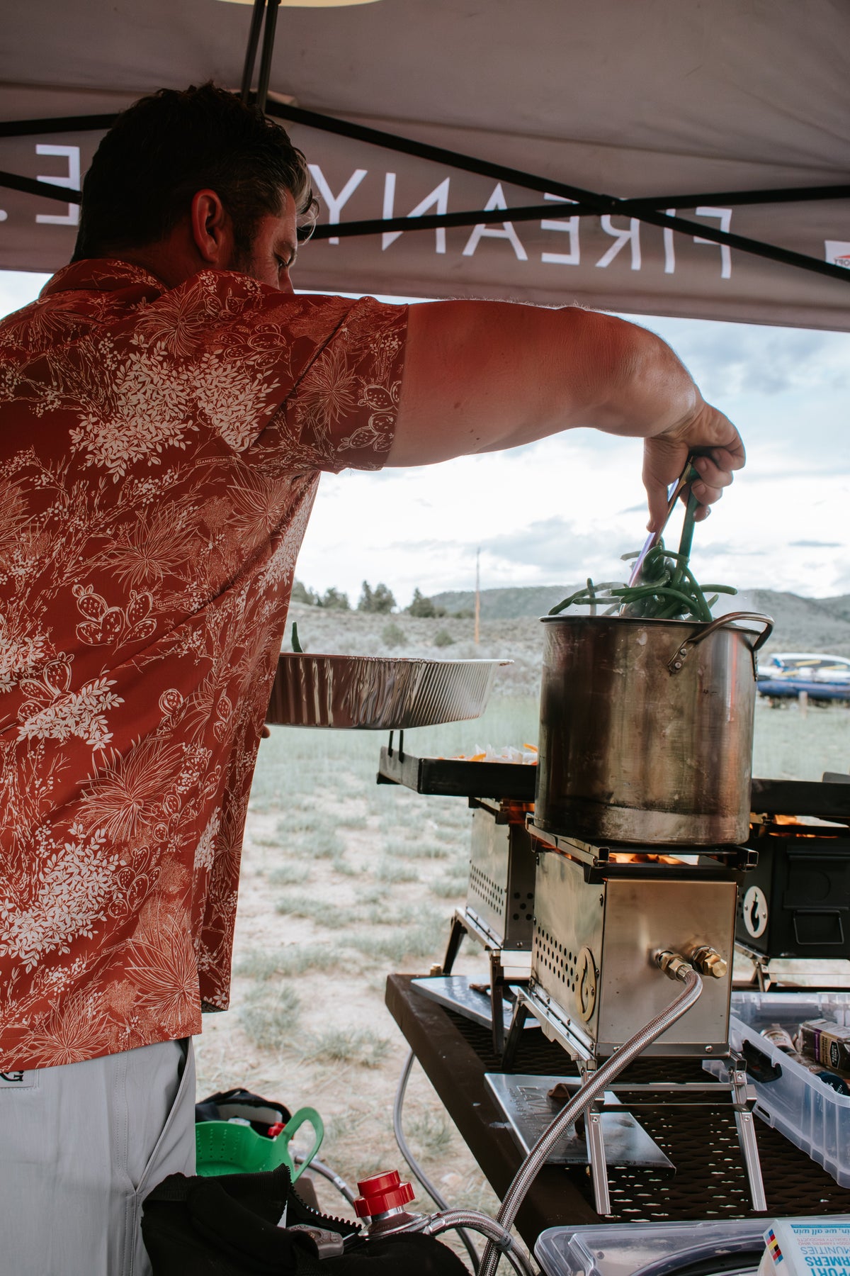 Outdoor event photo of a person tending a pot on a firebox setup under a branded canopy tent