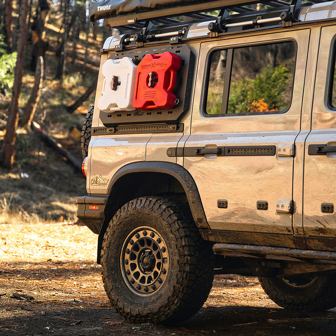 Rear side of a tan SUV with two fuel cans mounted on a black MOLLE panel in a wooded setting