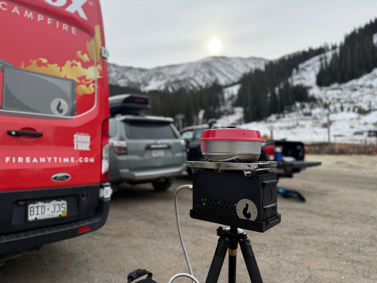 Outdoor daytime photo of a stainless firebox on its tripod stand at an event, with a branded van and mountain backdrop