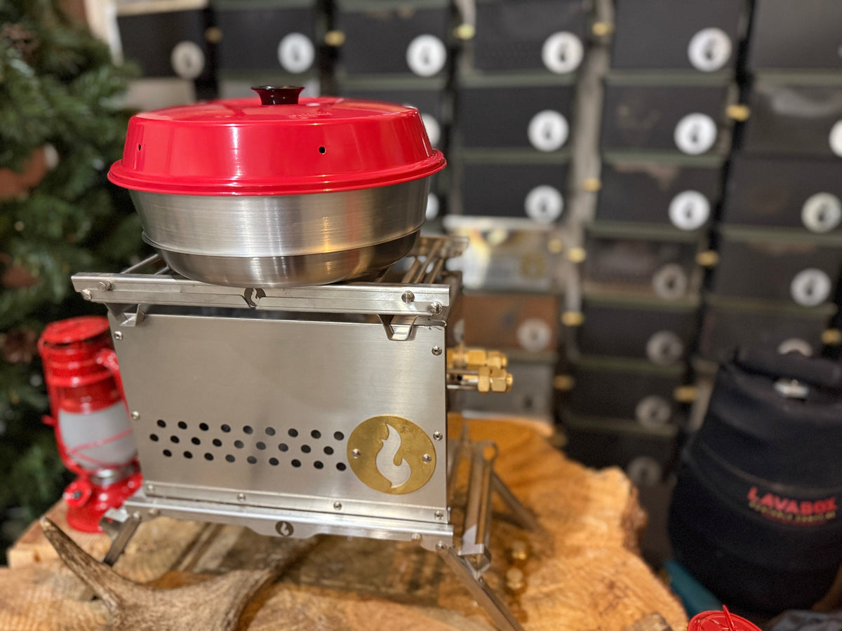 Close-up of the stainless steel Hekla stove topped with a mixing bowl and red lid, set on wood among pine branches with a lantern nearby