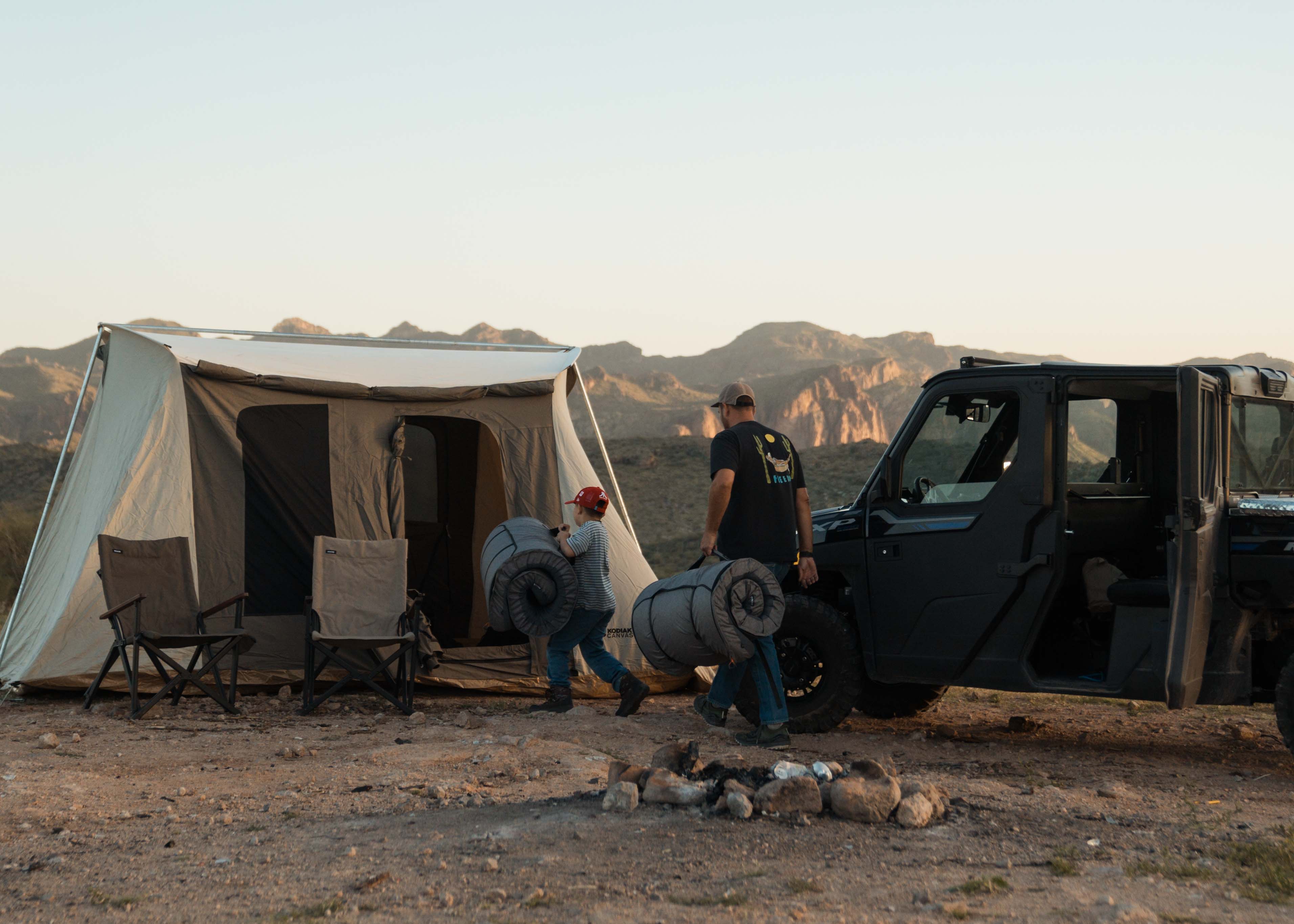 Two people setting up a large tent next to a black off-road vehicle in a desert landscape.