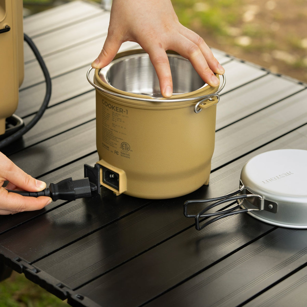 Hand holding the NESTOUT cooker's inner steel pot, showing the product label and the AC power inlet on the base with cord plugged in