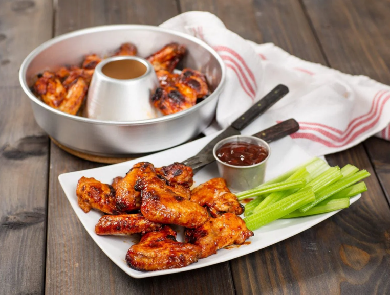 Plated chicken wings with celery and two containers of sauce on a wooden surface.