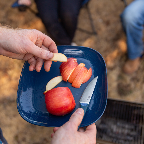 Hand placing a sliced apple piece onto a blue plate outdoors with the paring knife nearby