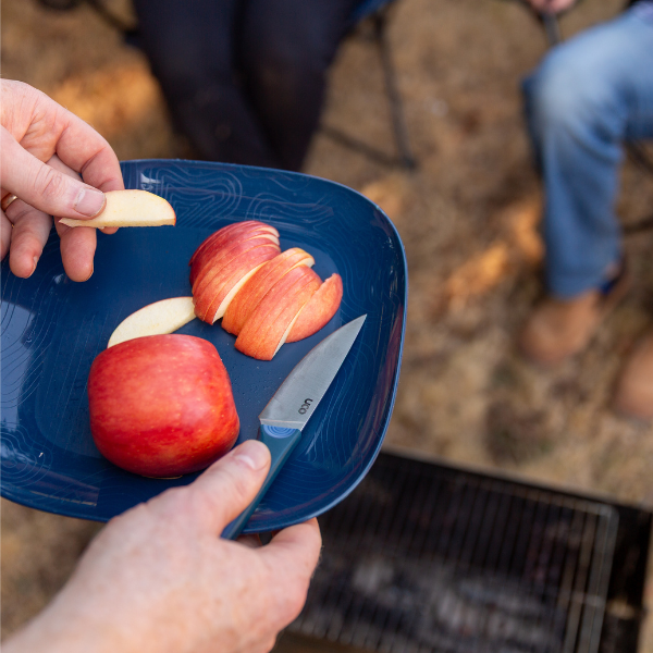 Hand placing a sliced apple onto a blue plate with a campfire visible in the background