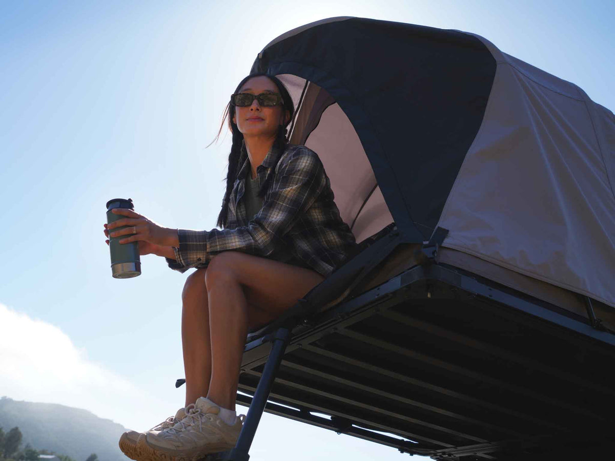 C6 Rev Rooftop Tent X (HellaStorm) – person seated at the platform edge holding a mug