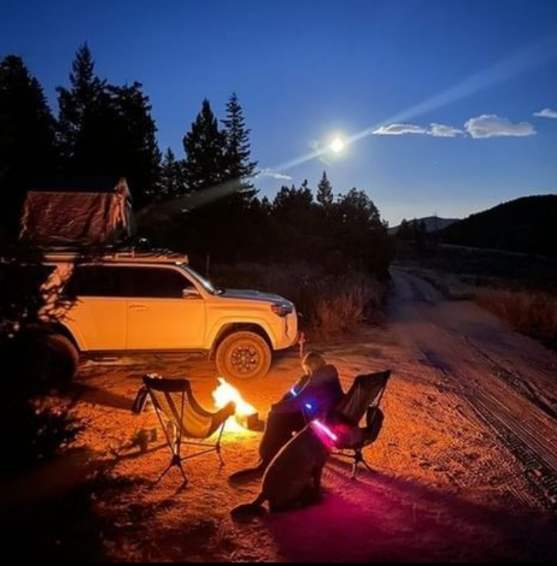 Nighttime lifestyle photo of a person next to a parked SUV and a lit fire pit under a starry sky with string lights nearby