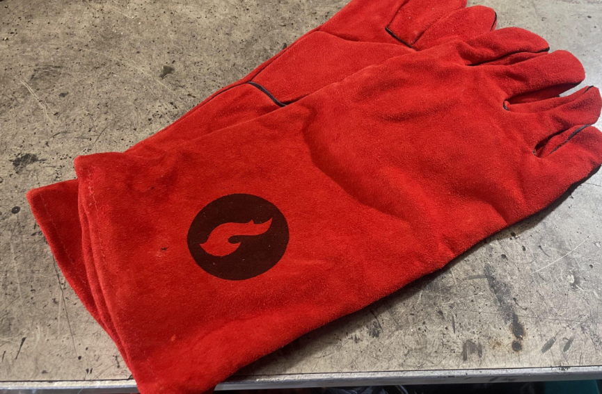 Pair of red leather fireproof gloves with a flame logo patch, resting on a concrete surface