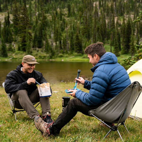 Two people seated outdoors near a lake and tent, eating from foil pouches with Switch Sporks