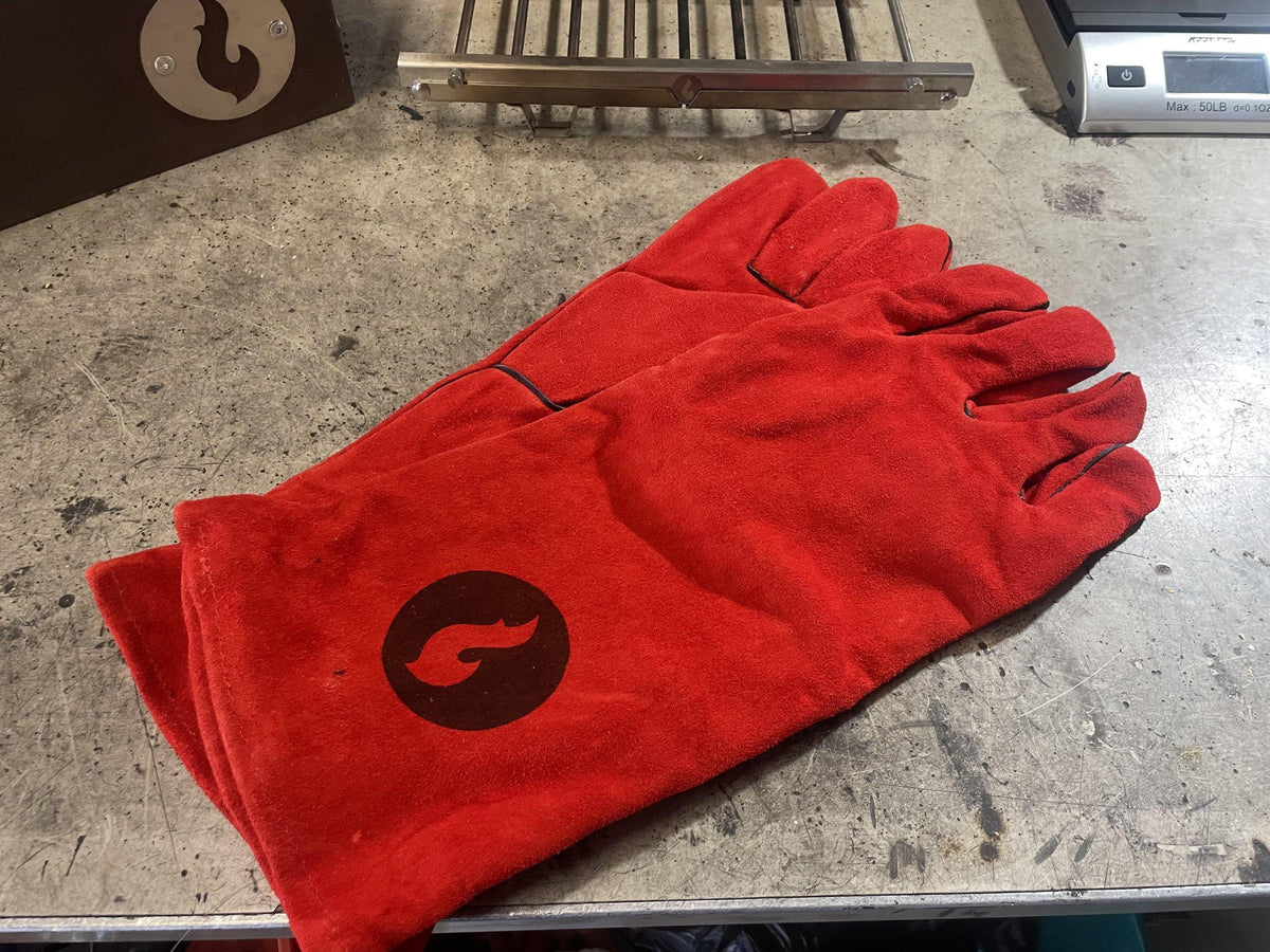Pair of red leather fireproof gloves with a flame logo patch, laid flat on a concrete workbench