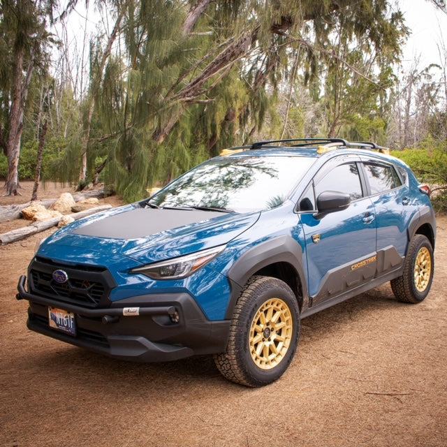 Blue Subaru SUV with gold wheels and a black front brush bar parked in a wooded area