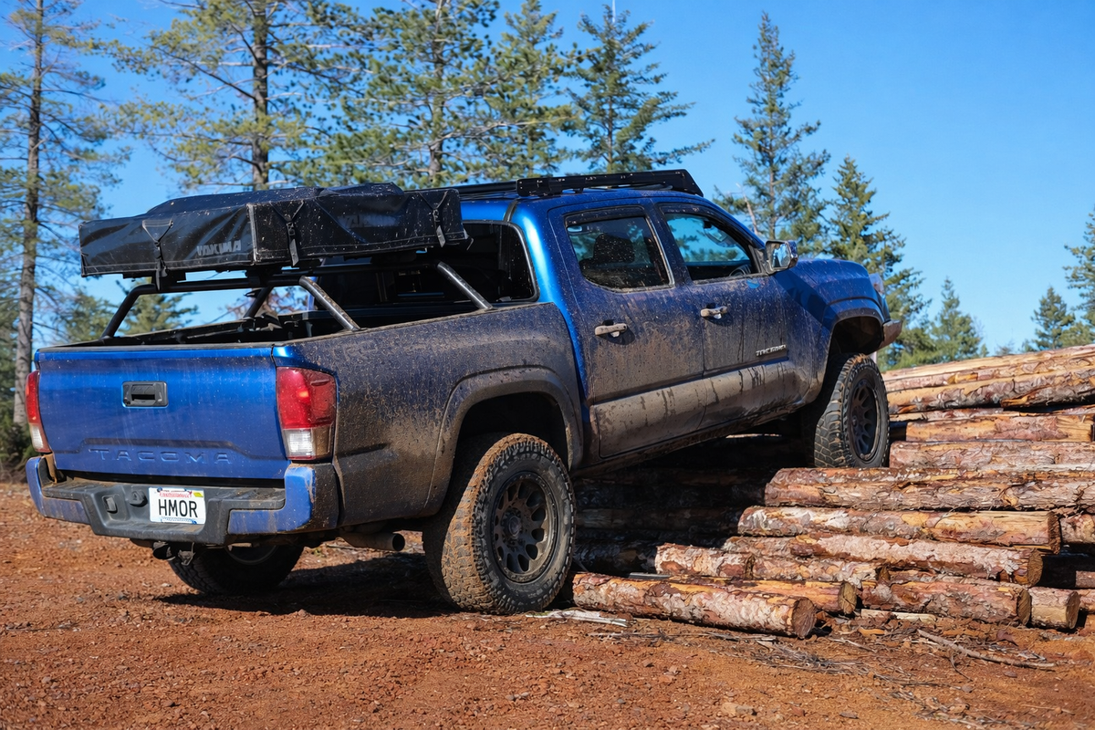 Blue Toyota Tacoma with HMOR bed bars and tent climbing a log obstacle