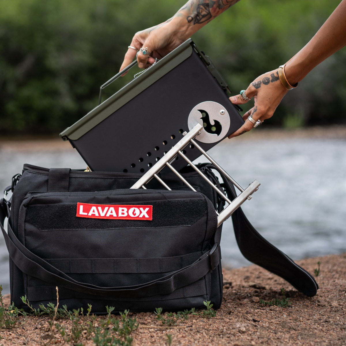Close-up of hands setting up a firebox next to its open LAVABOX carry bag near a riverbank