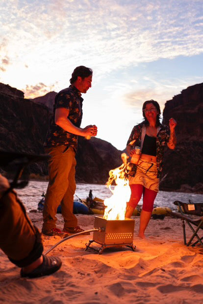 Lifestyle photo of two people warming their hands by a firebox burning on a sandy beach at dusk
