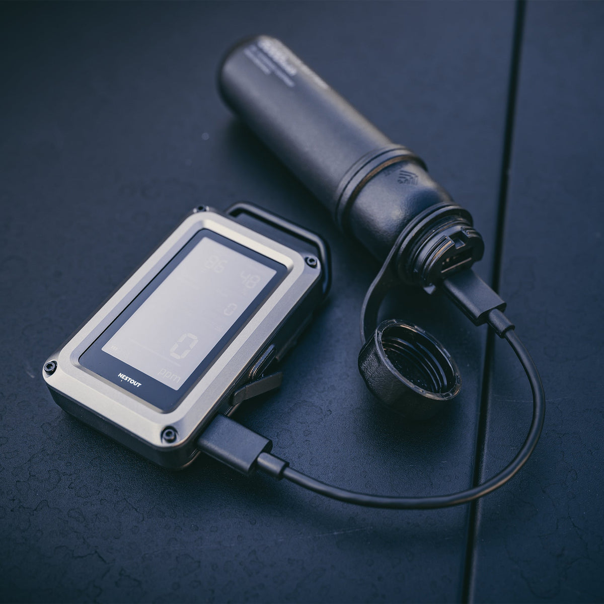 NESTOUT CO Alarm on a dark table connected by its USB-C cable to a black insulated bottle, screen showing temperature, humidity, and 0 ppm carbon monoxide