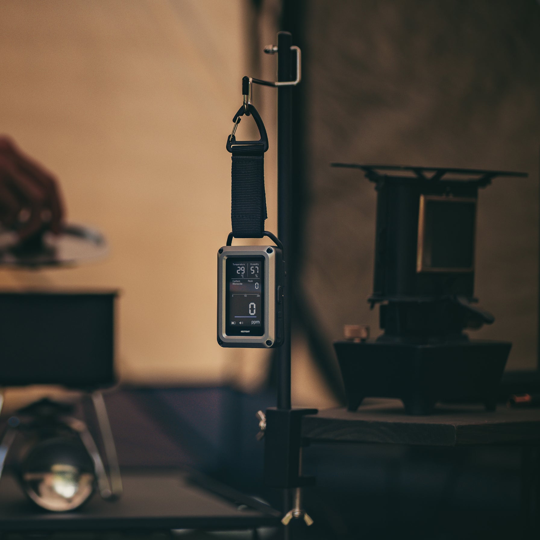 NESTOUT CO Alarm hanging by its strap and carabiner from a tent pole at a dim campsite, camp stove and lantern visible in the background