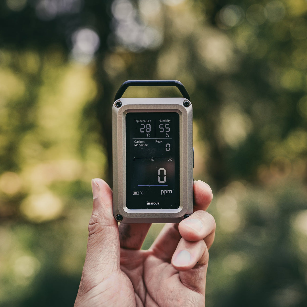 Hand holding the NESTOUT CO Alarm outdoors among green foliage, screen showing 28°C, 55% humidity, and 0 ppm carbon monoxide