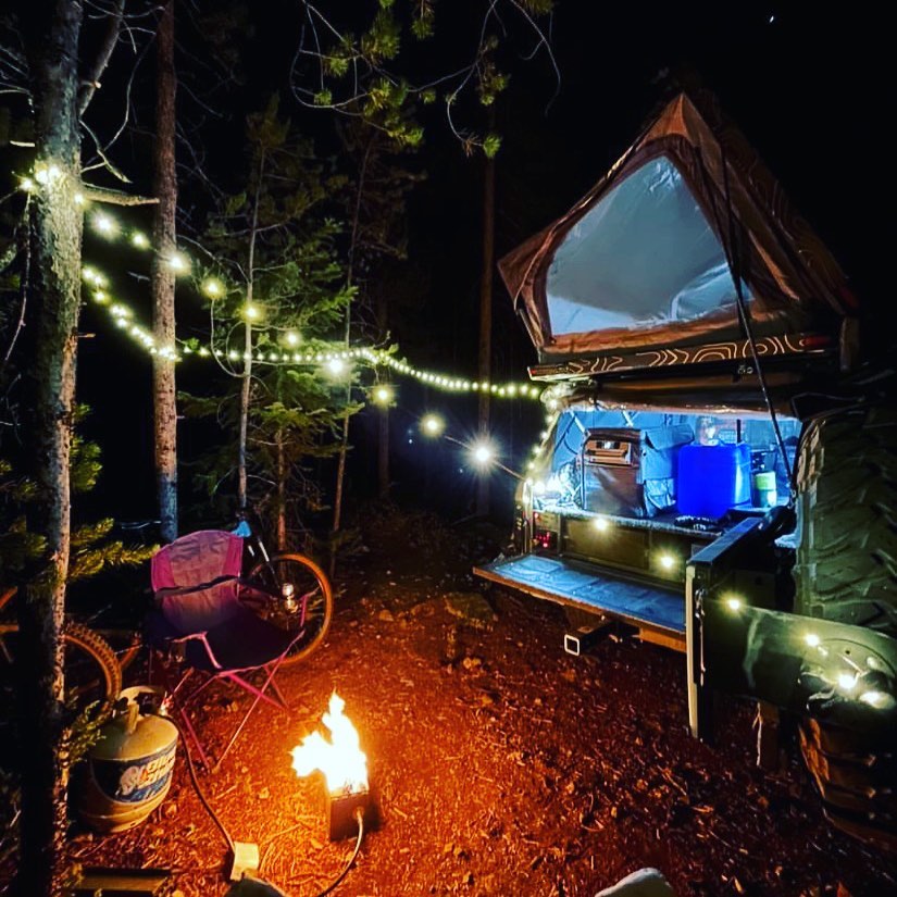 Nighttime campsite photo with string lights on a rooftop tent, camp chairs, and a lit fire pit