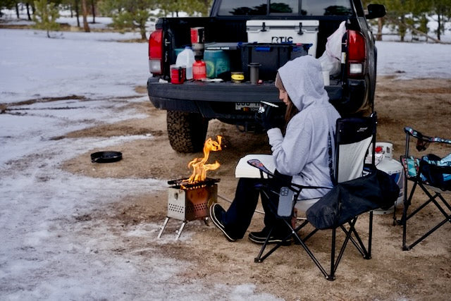 Lifestyle photo of a person seated in a camp chair next to a small firebox burning in the snow