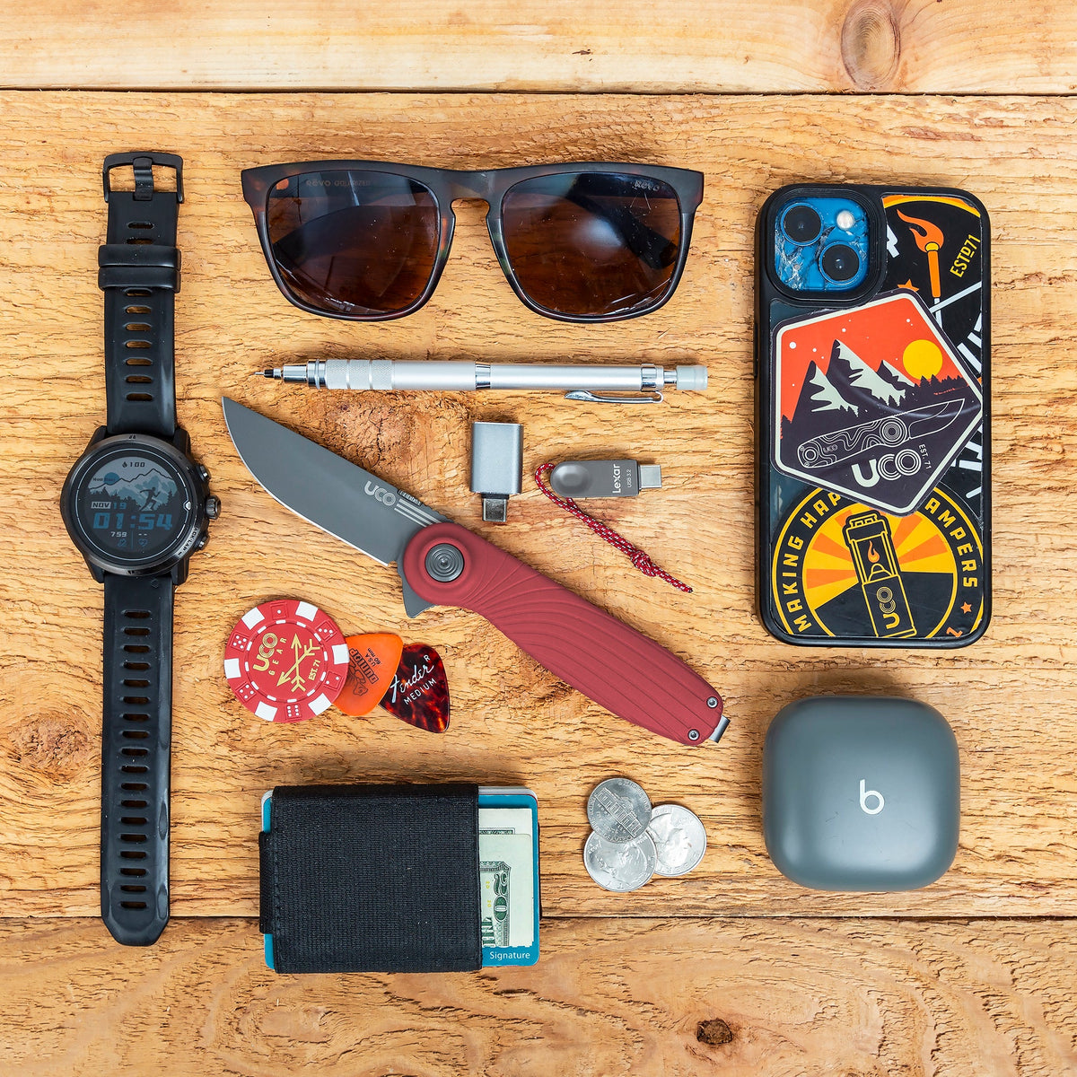 Flat lay of everyday carry gear including a watch, sunglasses, keys, a poker chip, and the red UCO Whidbey Mini folding knife