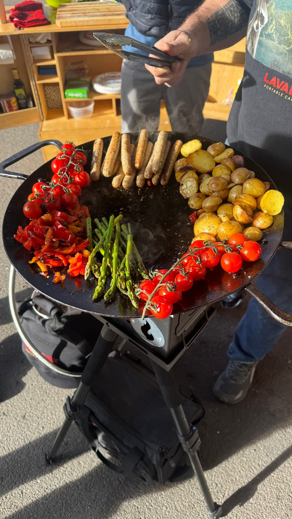 Overhead photo of vegetables and sausages grilling on a round steel griddle disc set over a firebox