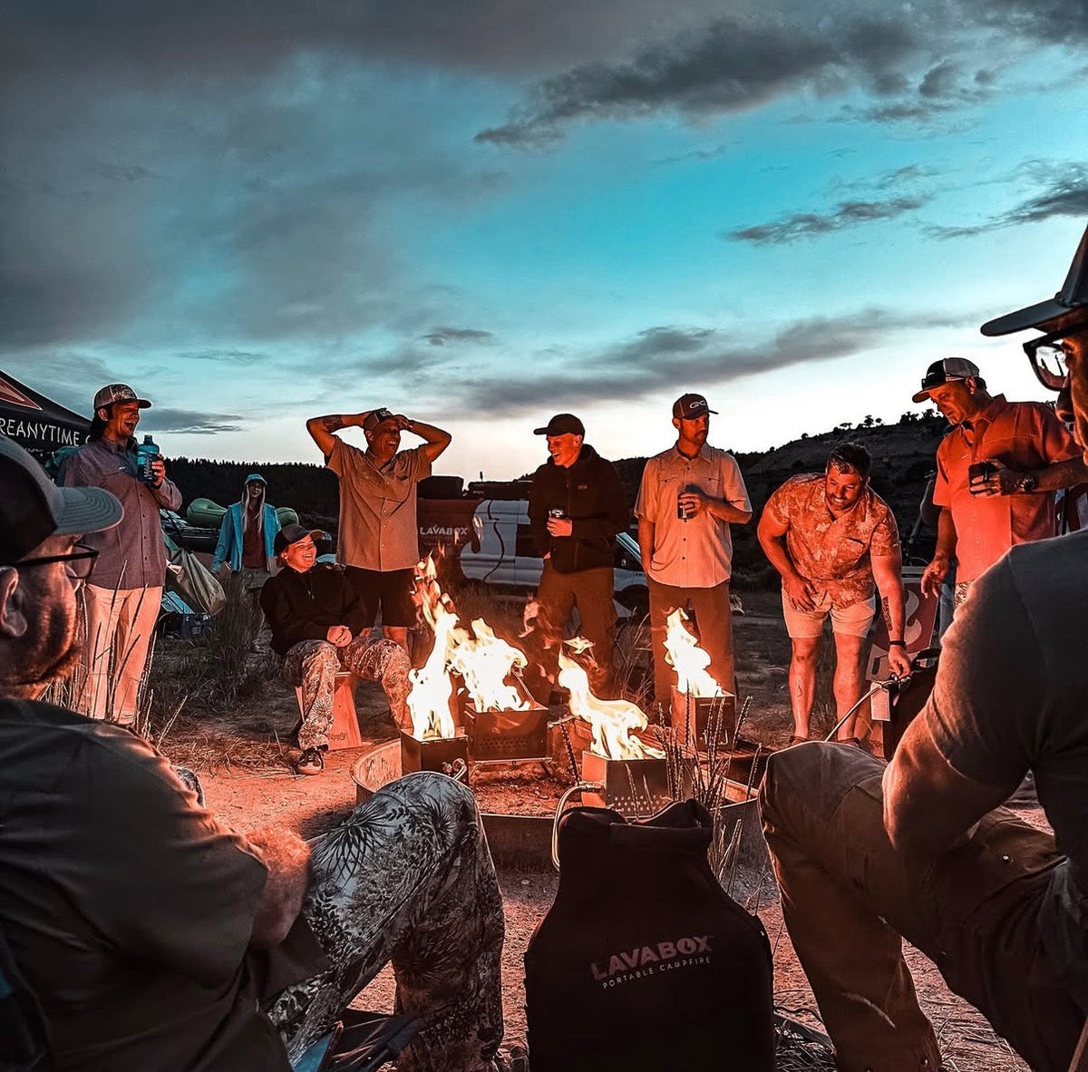 Nighttime lifestyle photo of a group gathered around multiple lit fireboxes at an outdoor gathering