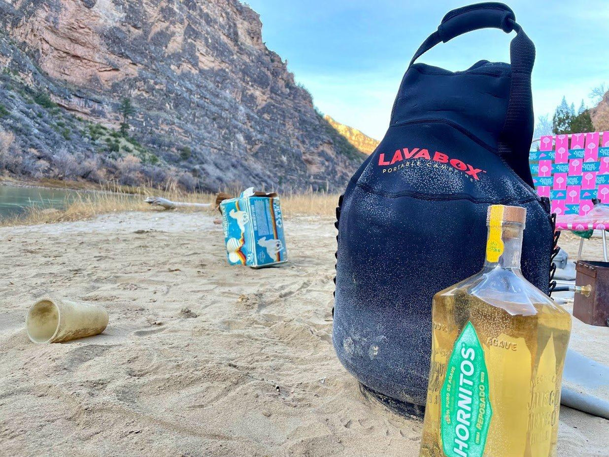 Lifestyle photo of a navy LAVABOX tank top propane cover and a fuel container on a sandy riverbank