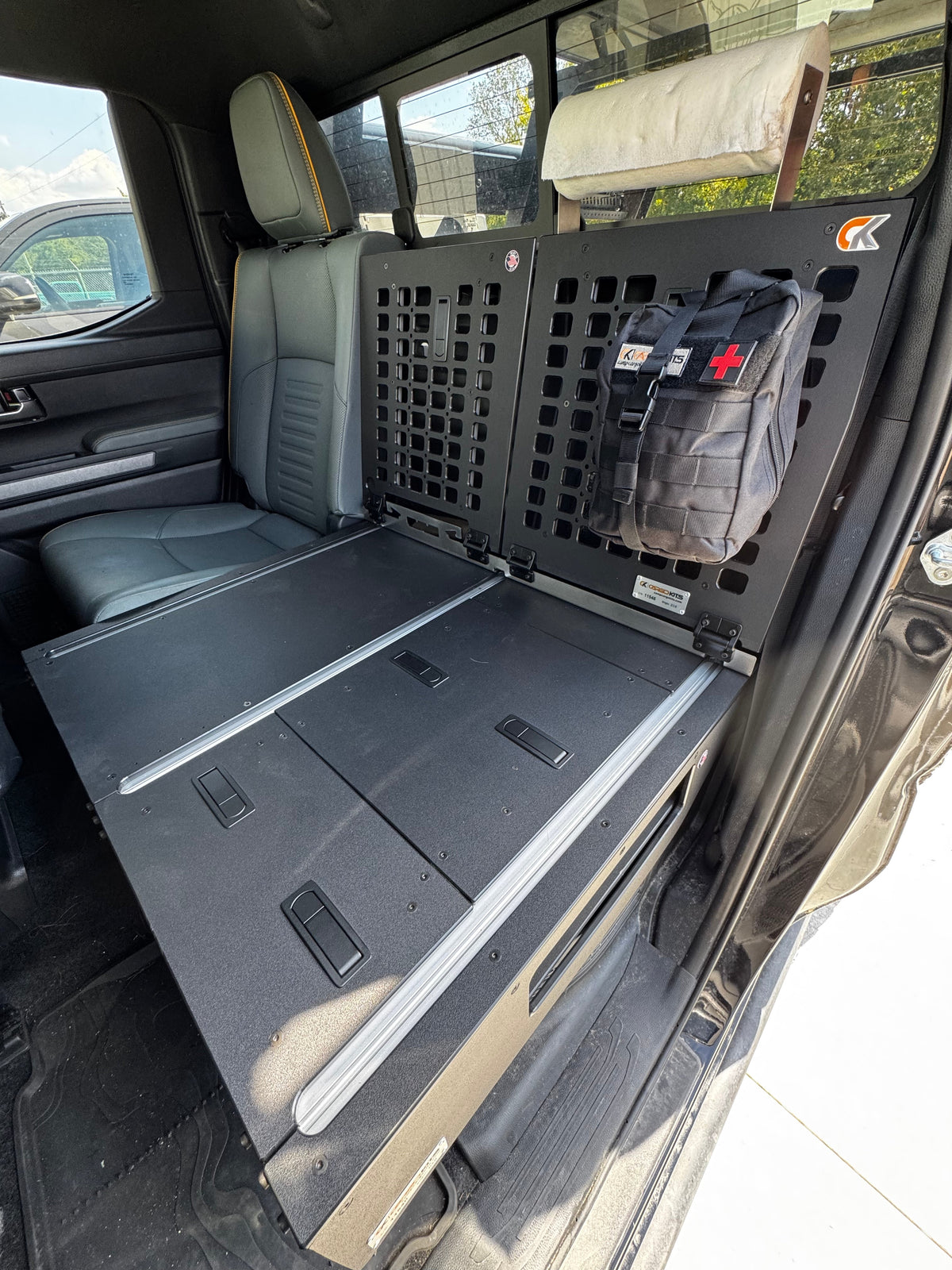 Angled view of the CargoKits seat delete platform showing hatch lids, silver accessory rails, and the perforated MOLLE rear panel with a first-aid pouch and paper towel roll