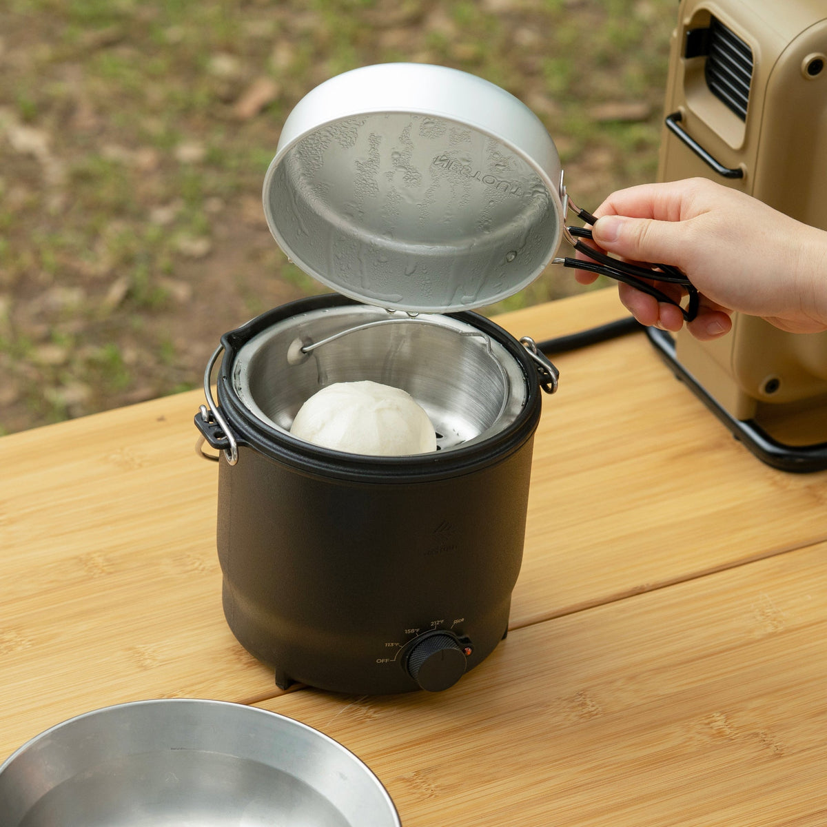 Lifting the lid off the NESTOUT cooker to reveal a steamed bun on the steaming rack inside, with the temperature dial visible on the unit