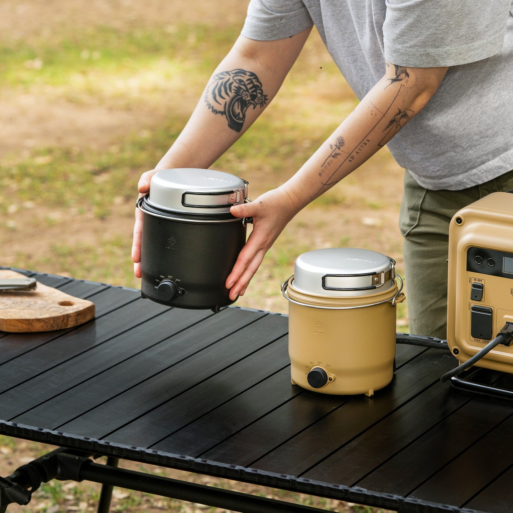 Holding the black NESTOUT cooker beside a tan NESTOUT cooker on a table next to a power station