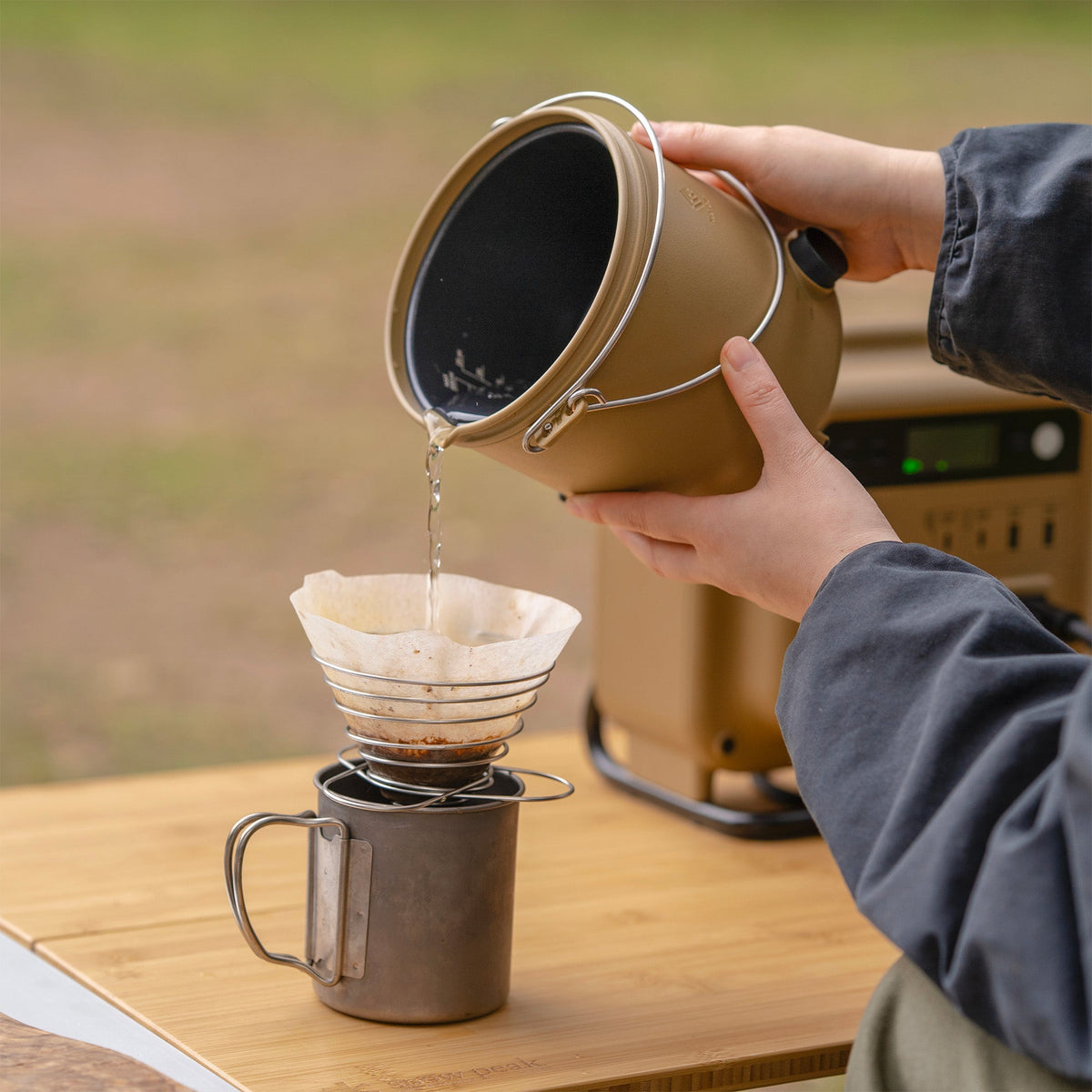 Pouring hot water from the NESTOUT cooker's pot into a pour-over coffee filter set over a titanium camp mug, with a power station in the background
