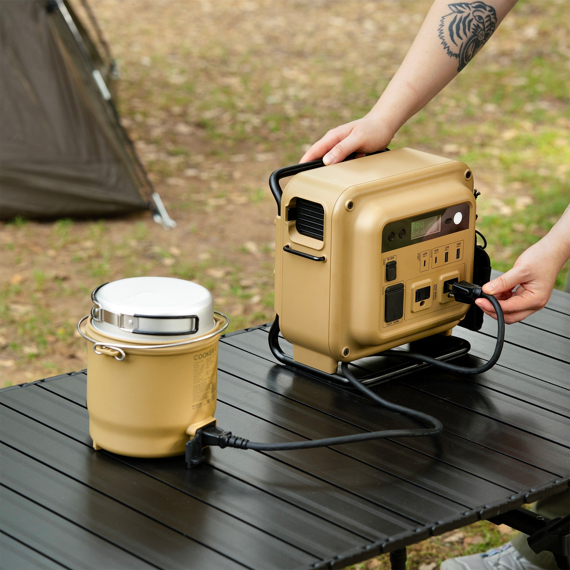 Hand plugging the NESTOUT cooker's power cord into a NESTOUT portable power station on a picnic table, tent visible in the background