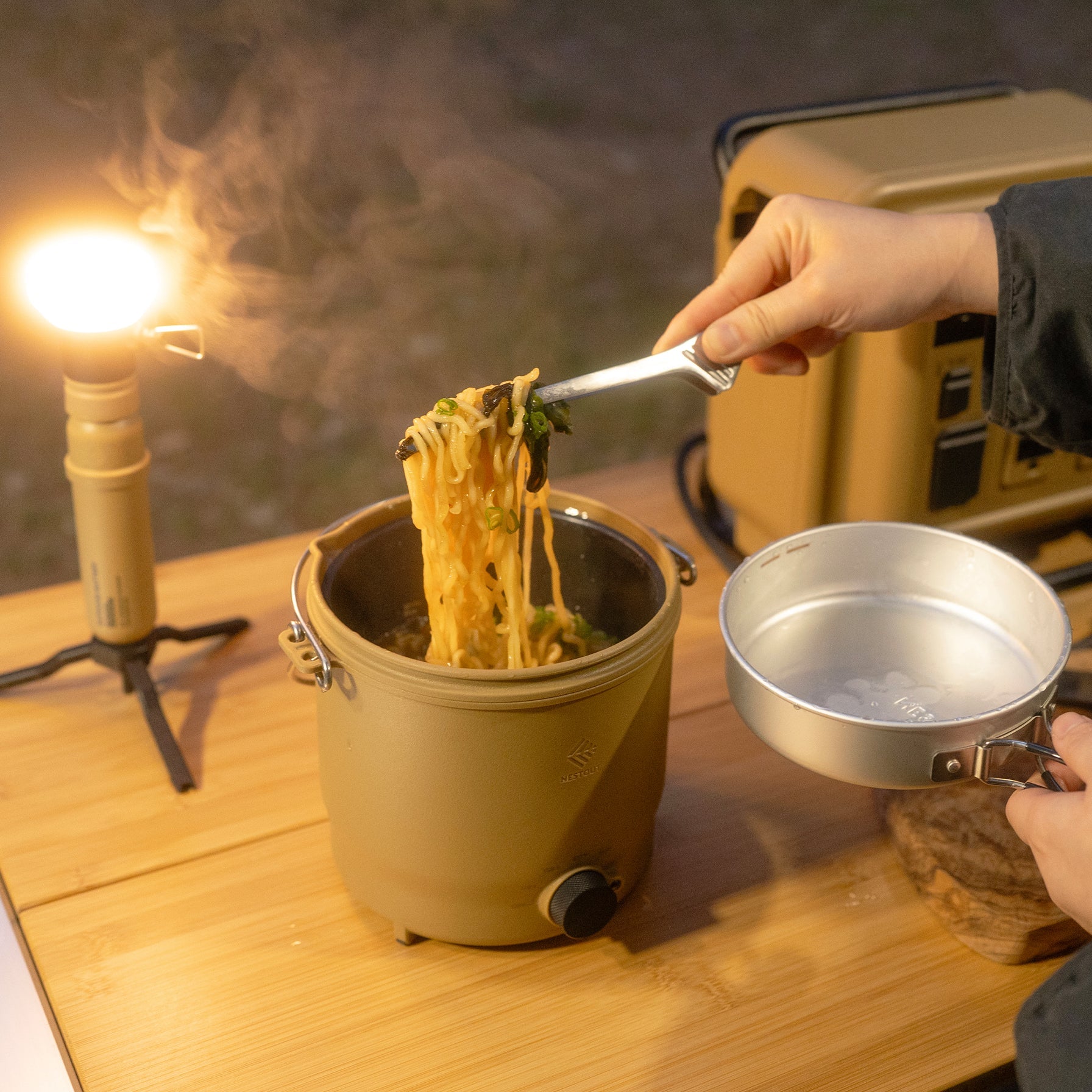 Using tongs to lift ramen noodles from the NESTOUT cooker's pot near a glowing lantern at a nighttime campsite