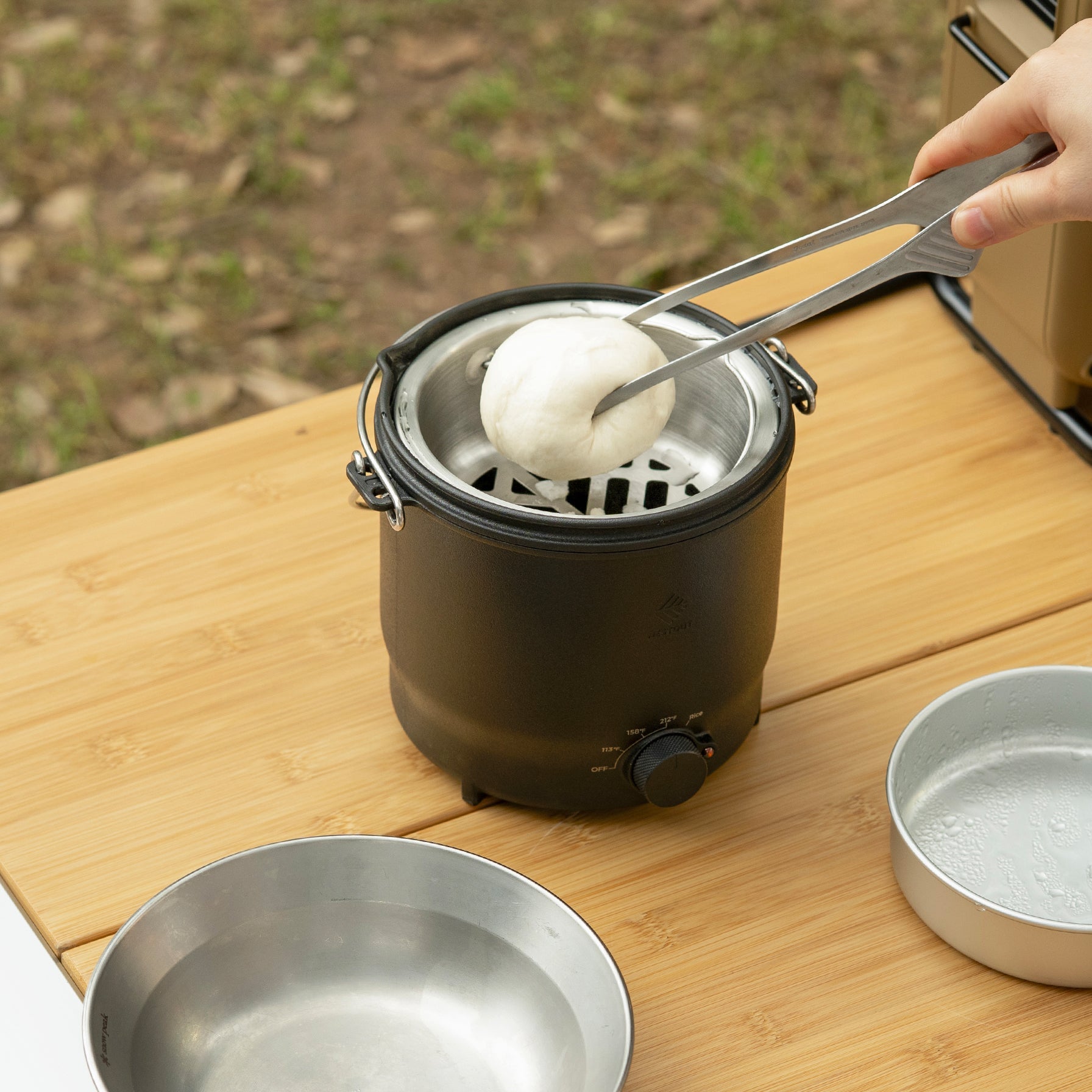 Lifting a steamed bun from the NESTOUT cooker's steaming insert with tongs, the condensation-beaded lid resting beside the pot