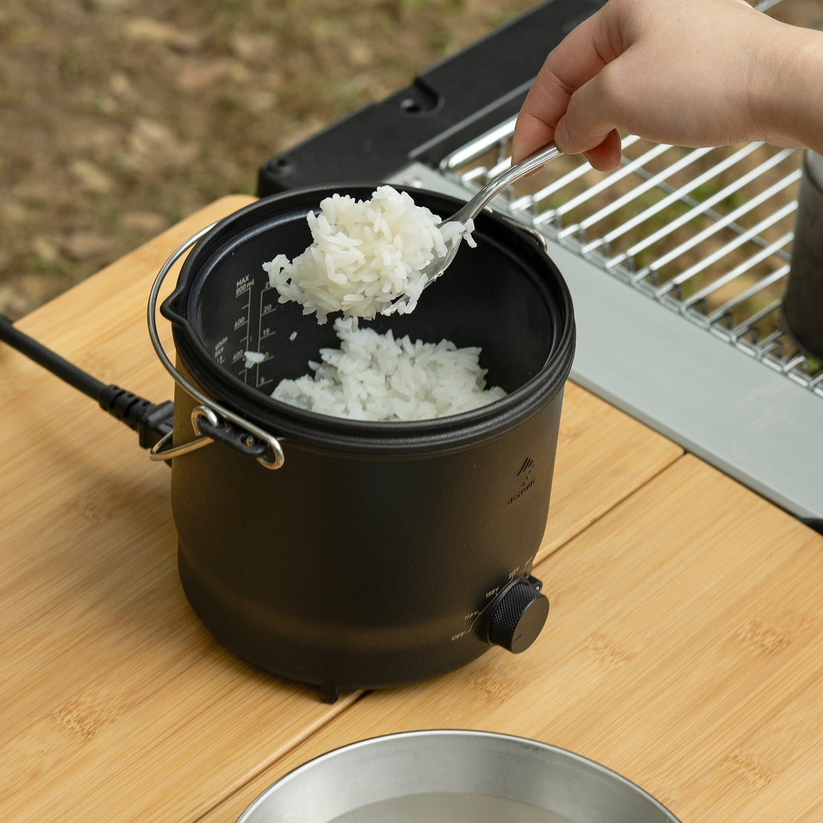 Scooping cooked white rice from the NESTOUT cooker with a spoon at a campsite, a portable grill visible in the background