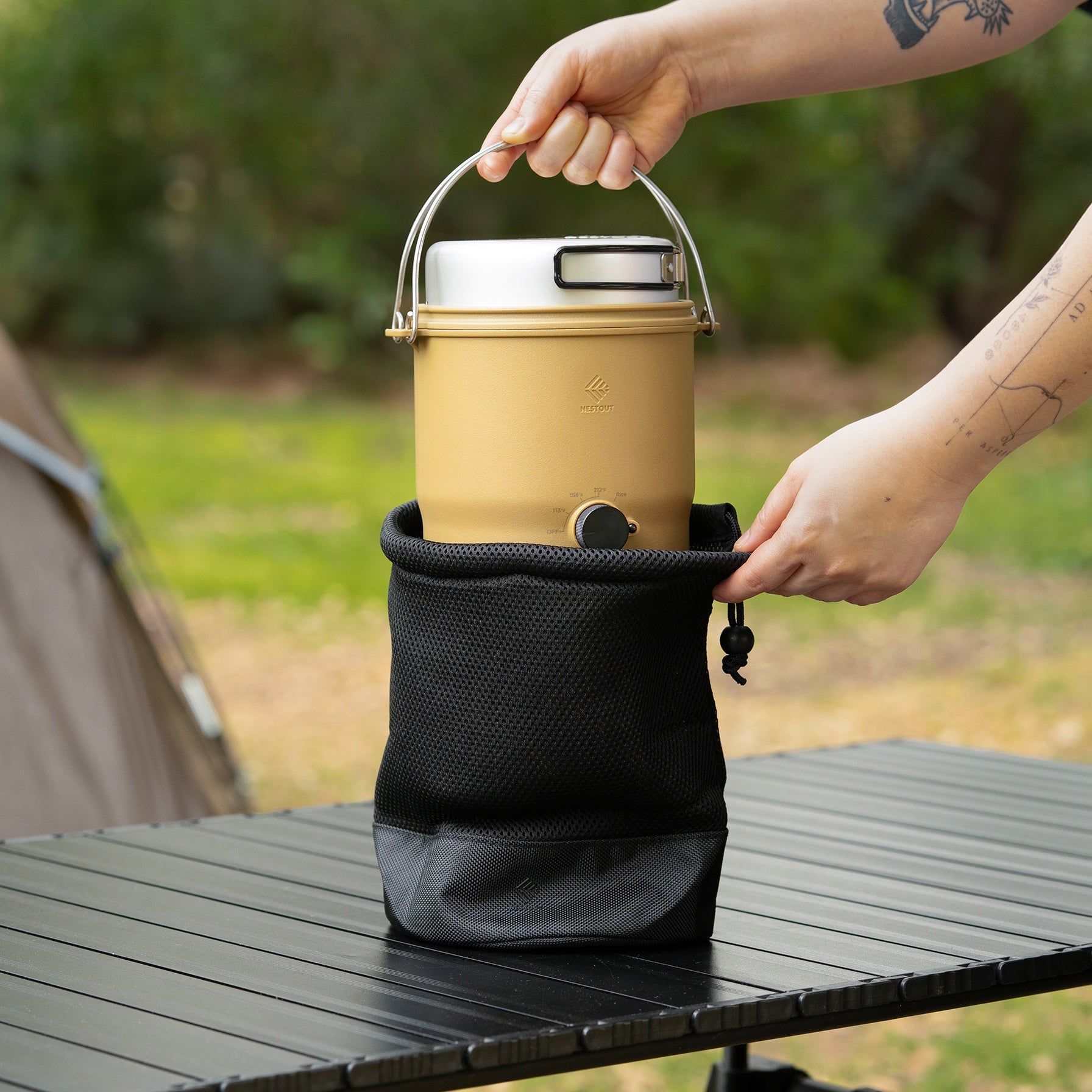 Lifting the assembled beige NESTOUT cooker out of its black mesh storage bag near a tent
