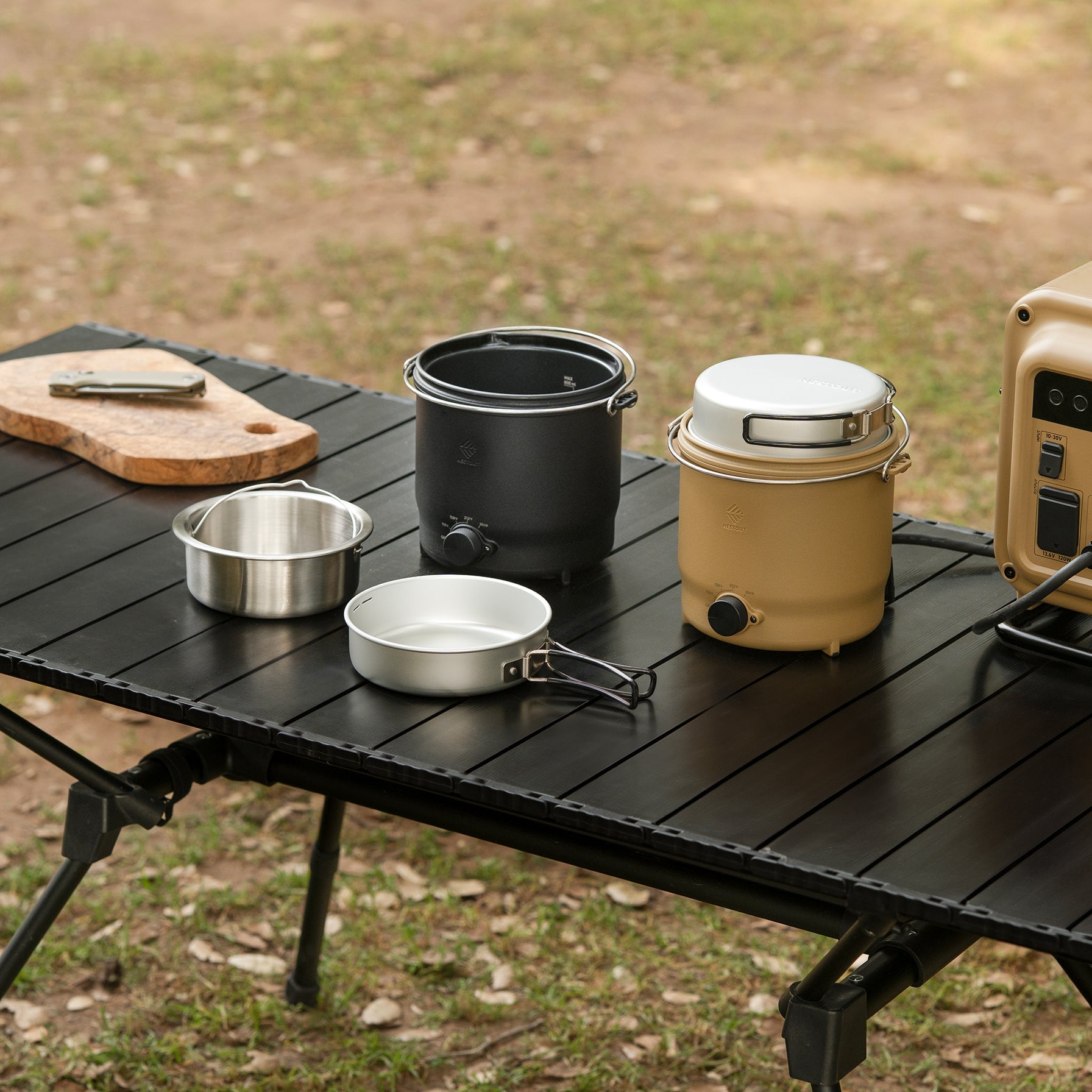 Overhead view of a black NESTOUT cooker (open, empty pot) beside a closed beige NESTOUT cooker on a picnic table, with the steaming insert, a small lid piece, and a power station nearby