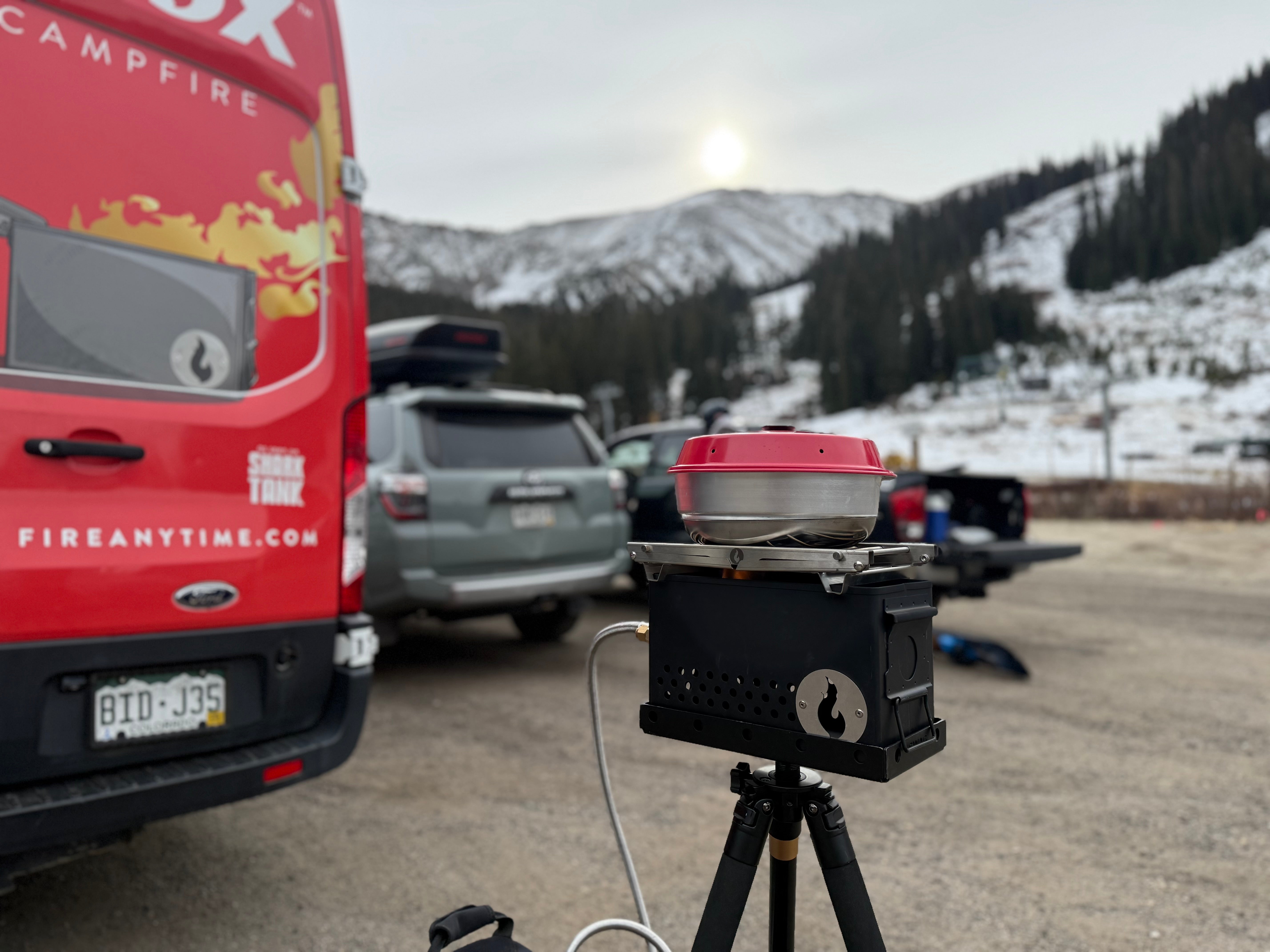 Red and black Omnia camping stove on a tripod with a scenic mountain backdrop