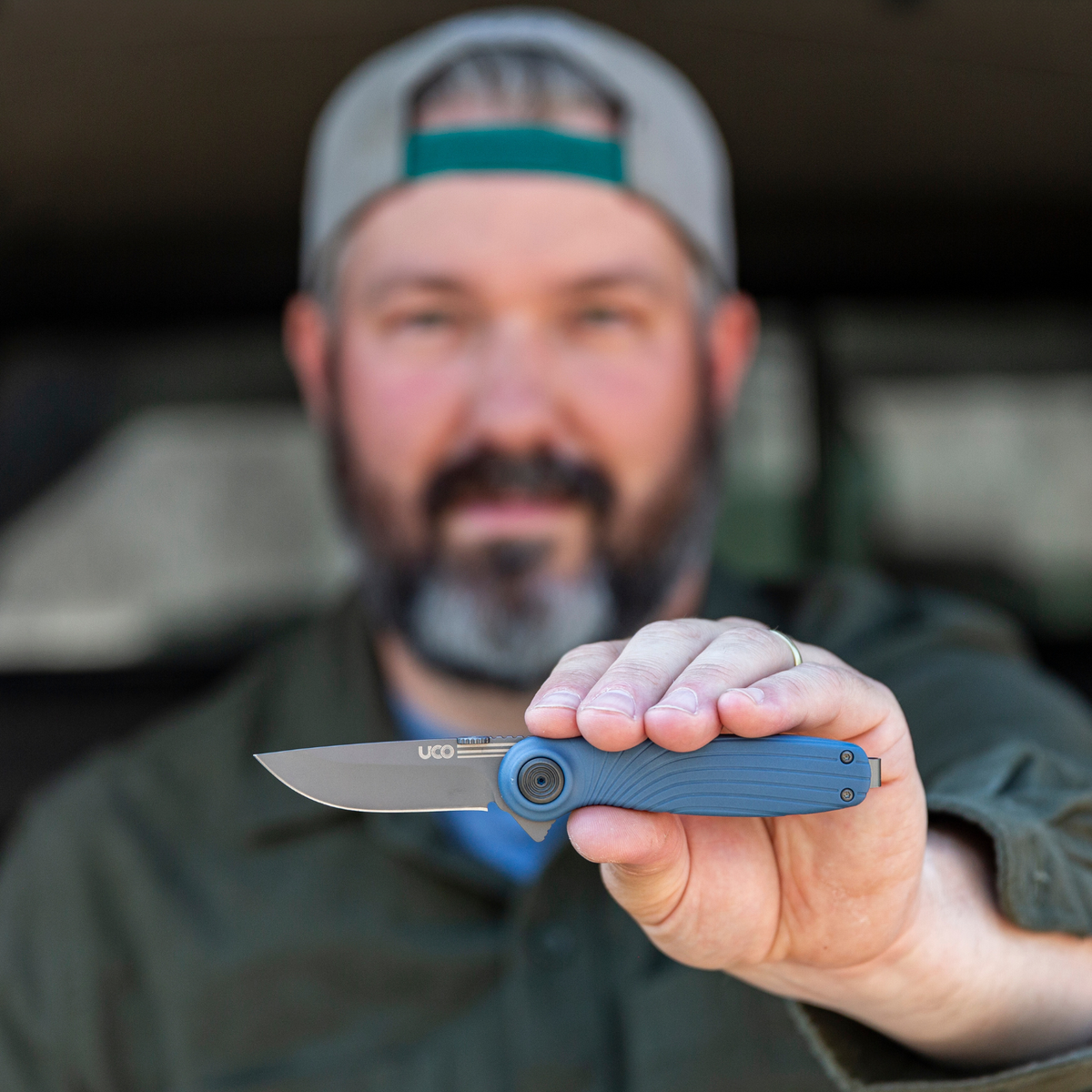 Man in a ballcap holding a navy blue UCO Whidbey Mini folding knife near a truck