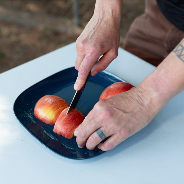 Hand slicing an apple with the UCO Camp Paring Knife on a blue plate, alternate angle