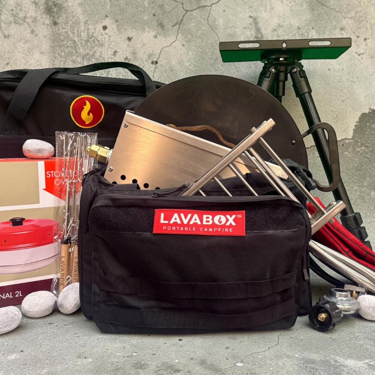 Overhead flat-lay of a full kit with griddle disc, tripod stand, roasting forks, Hades Stones, and carry bag arranged on a concrete surface