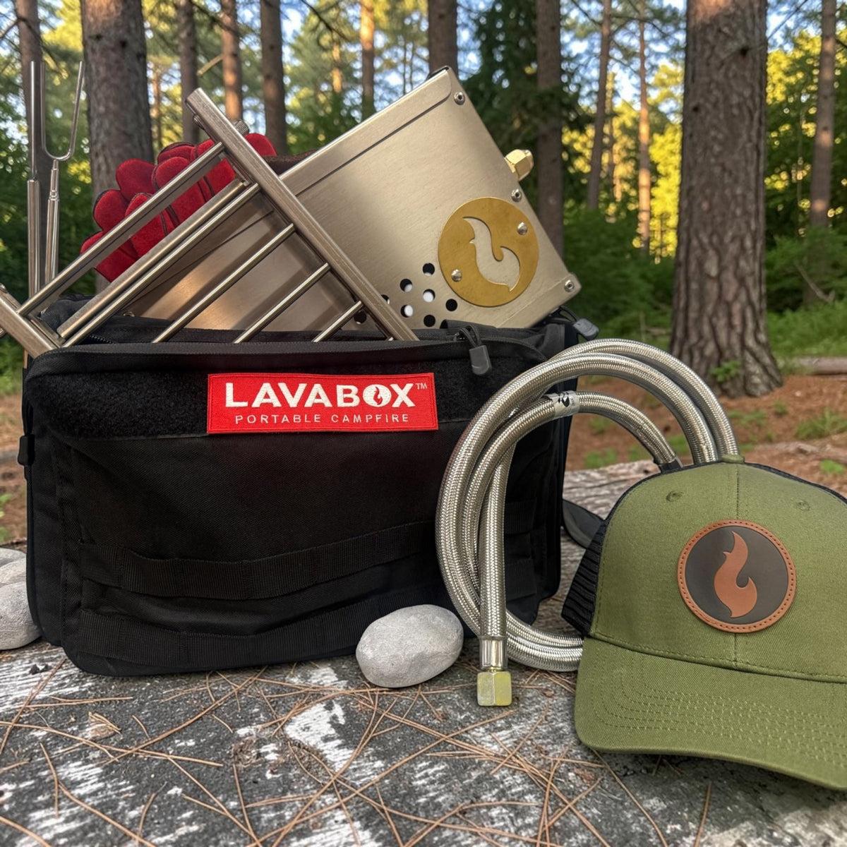 Overhead flat-lay of a stainless firebox kit with hose, roasting forks, gloves, a cap, and Hades Stones next to an open LAVABOX carry bag