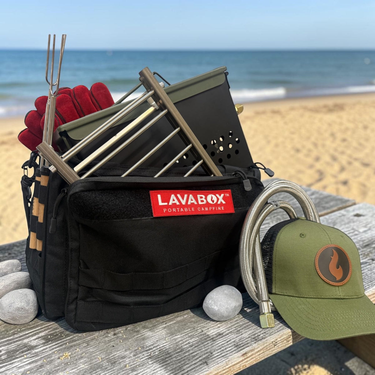 Outdoor beach photo of a packed LavaBox kit with firebox, roasting forks, gloves, hose, and a cap arranged on a picnic table by the shore