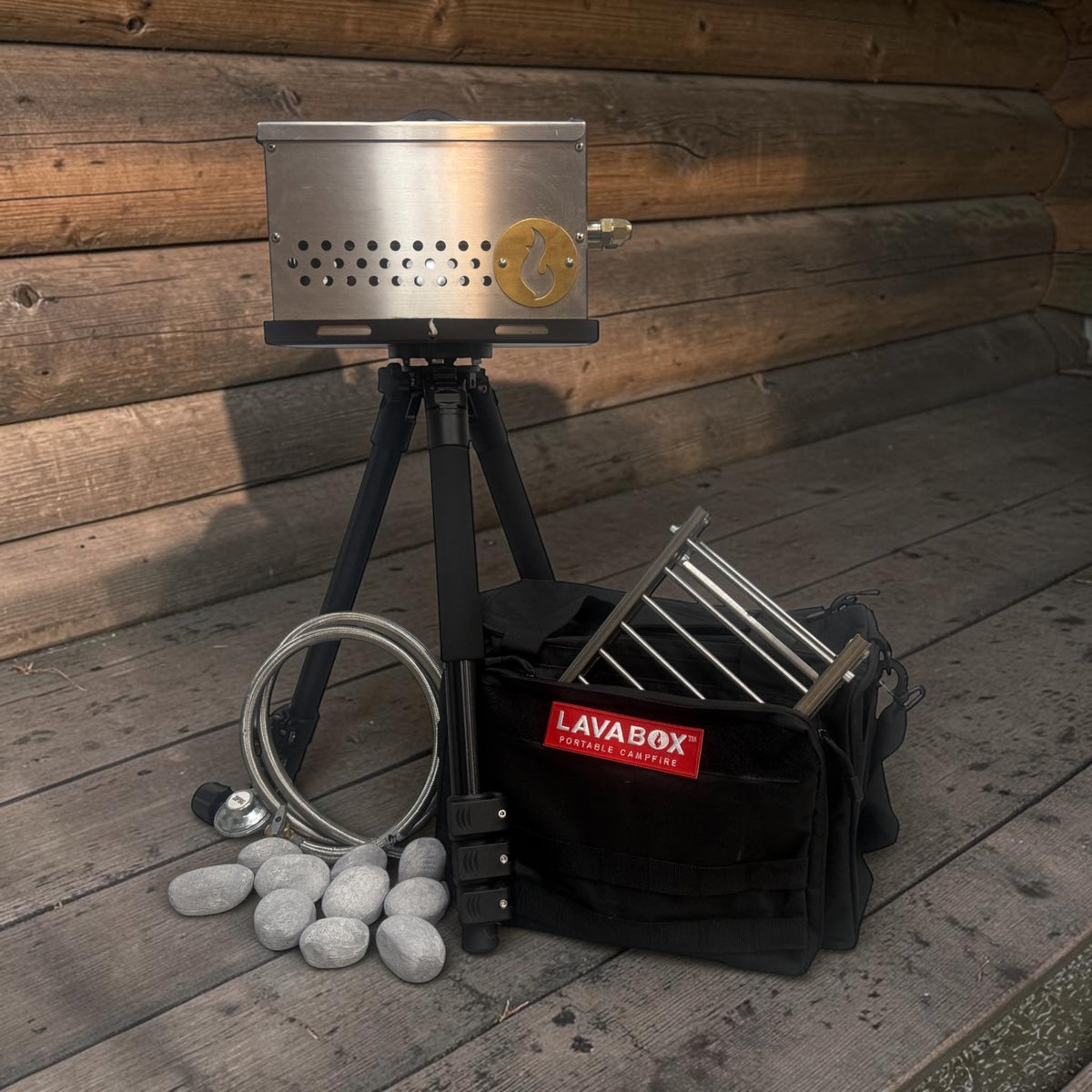 Outdoor photo of a stainless firebox on its tripod stand on a wood deck, with a coiled hose, roasting forks, and its LAVABOX carry bag alongside