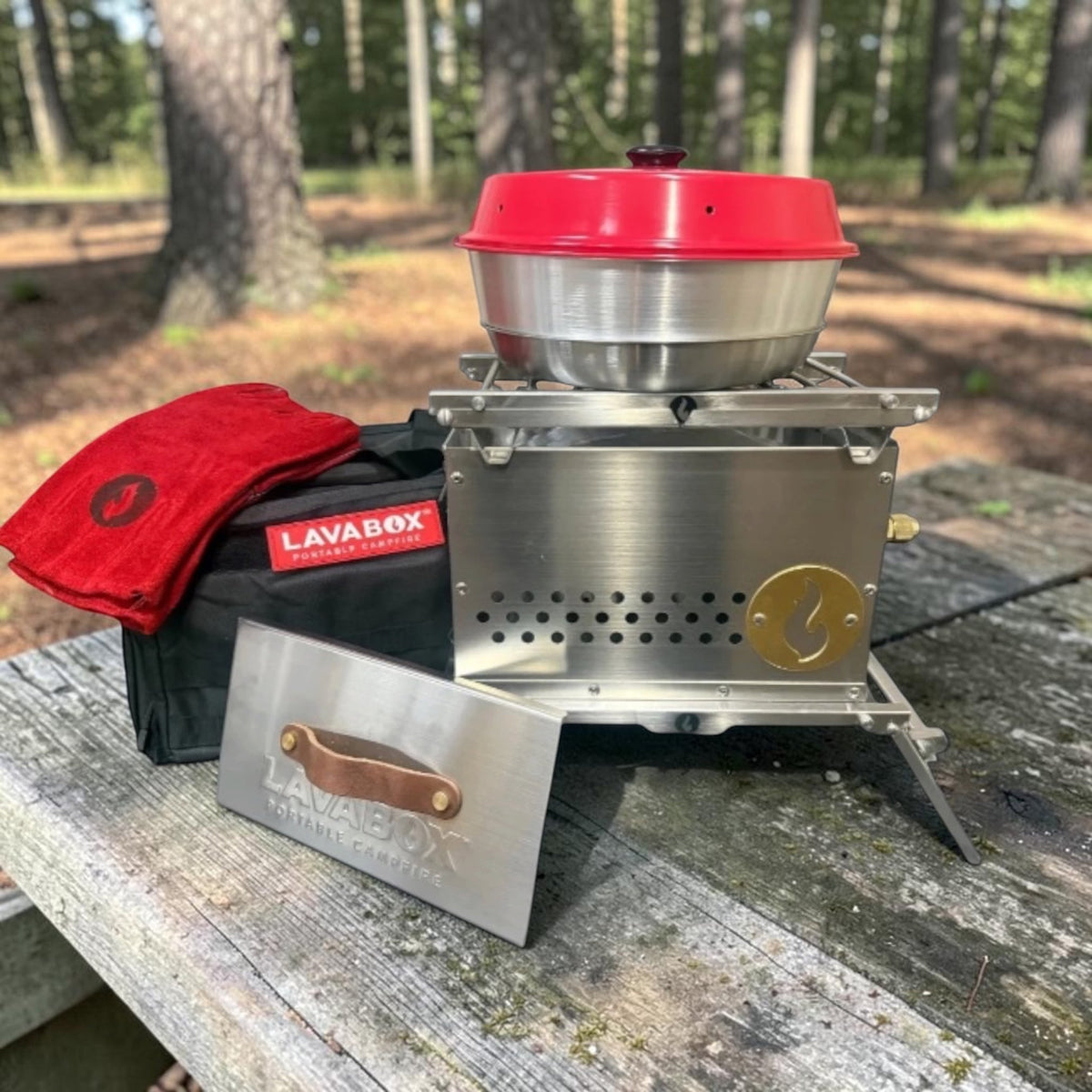 Outdoor photo of the stainless steel Hekla stove topped with a mixing bowl and red lid, resting on a picnic table with its case and roasting fork alongside