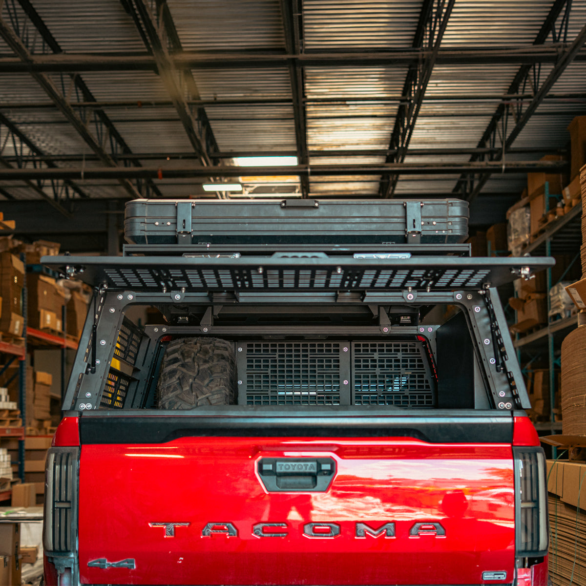 Rear hatch closed on a bed rack on a red truck, with a roof-mounted case and a fuel can mounted on the panel