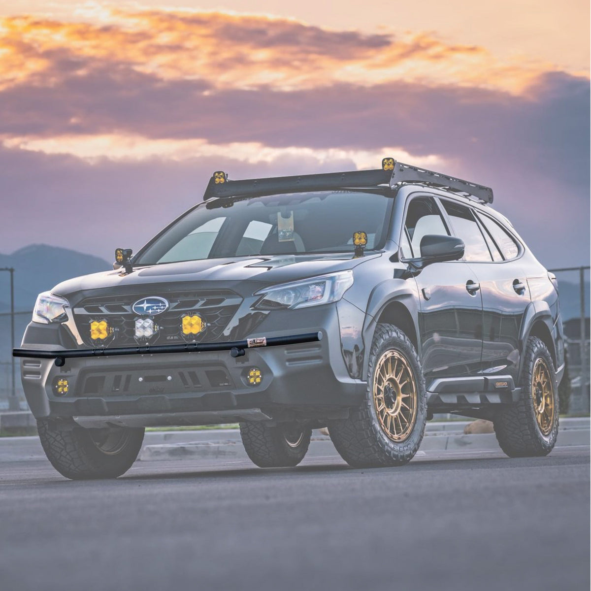 Dark Subaru wagon with gold wheels and amber lights on a front brush bar at sunset